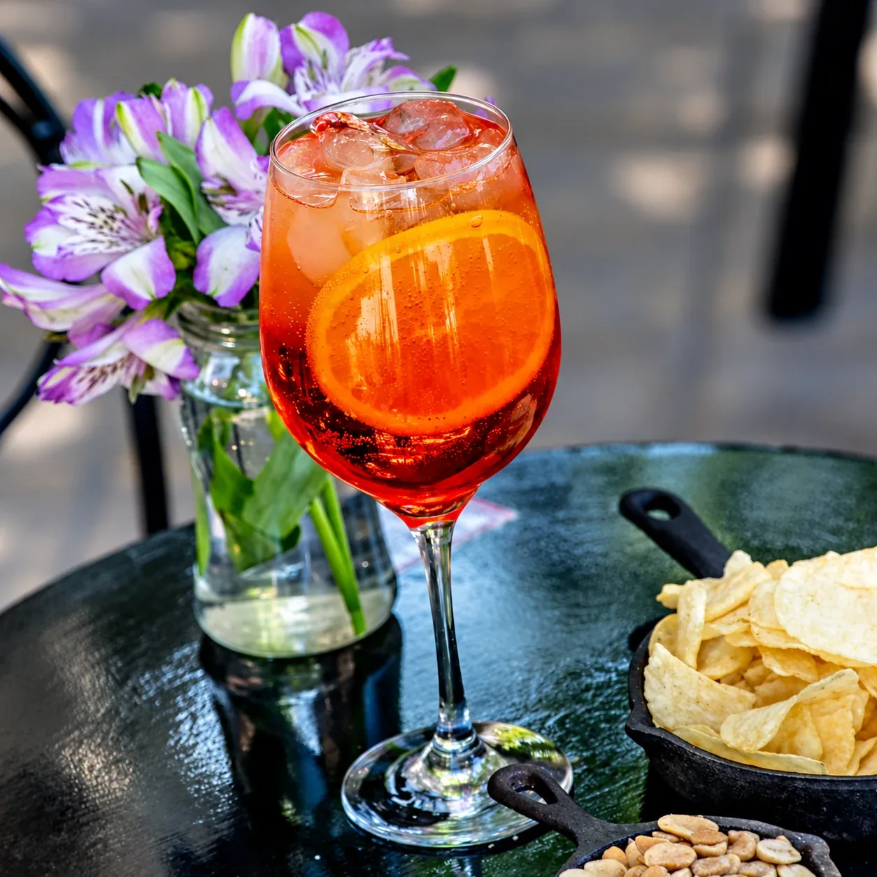 Un spritz con una rodaja de naranja, con papas fritas y maní, en una mesa de la vereda